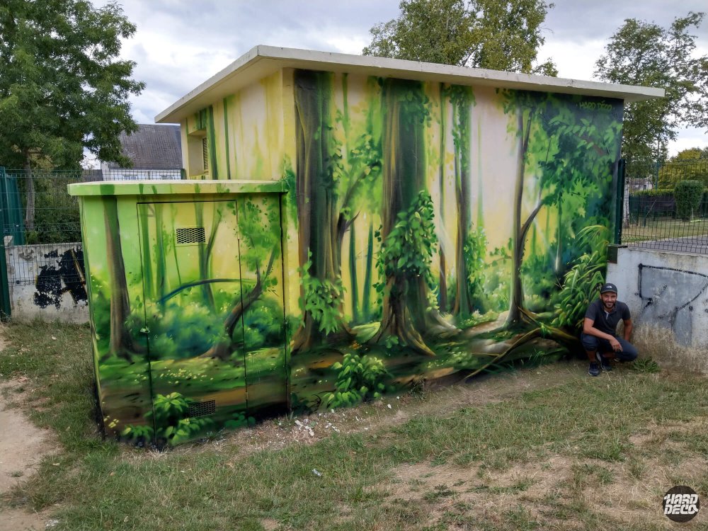 Artiste posant devant la fresque forêt sur transformateur EDF, vue face arrière avec troncs et feuillages aérosol