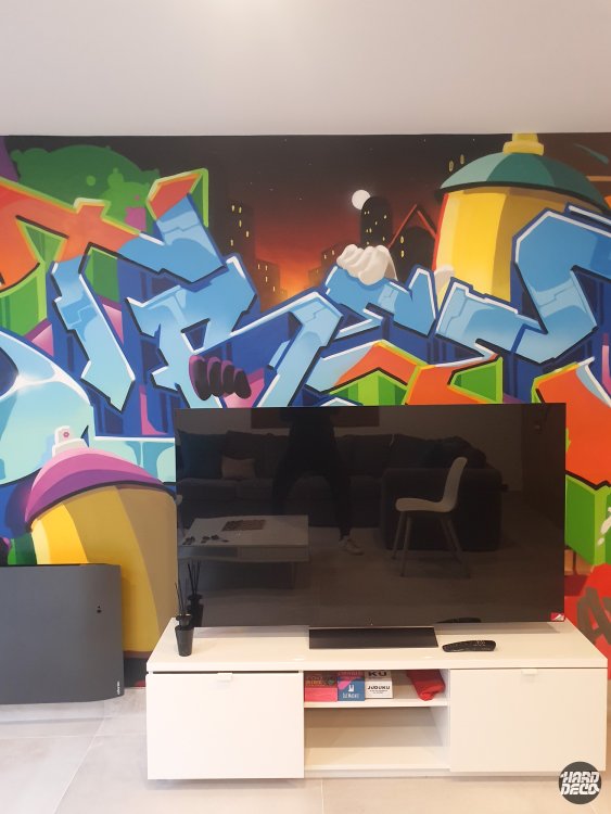 Fresque graffiti salon, plan rapproché sur les lettres bleues glacées et la skyline nocturne, derrière la télévision