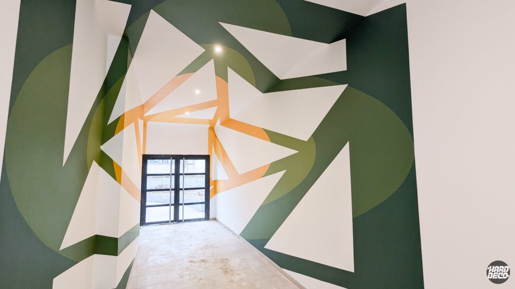 Anamorphose géométrique en vert forêt et ocre doré couvrant murs et plafond d'un hall de résidence, vue en perspective latérale