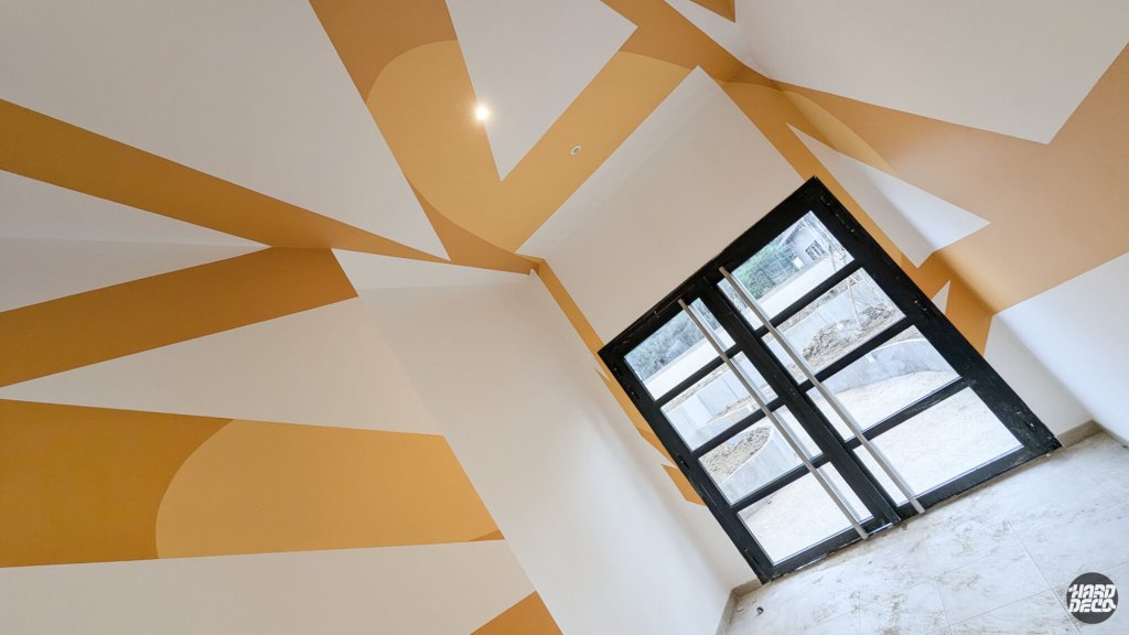 Détail du plafond peint en formes géométriques ocre et blanc dans le hall d'entrée de la résidence SogeProm, vue en contre-plongée