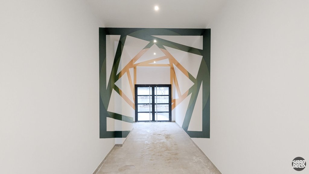 Vue frontale de l'anamorphose géométrique dans le couloir de la résidence SogeProm, composition triangulaire verte et ocre sur murs et plafond