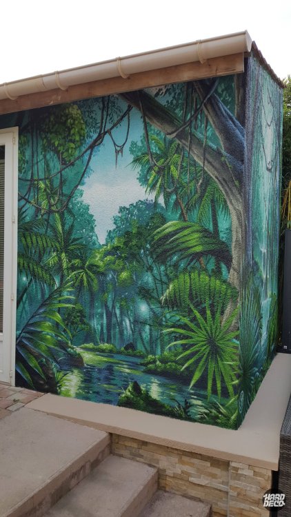 Fresque jungle aérosol sur angle de façade extérieure, végétation tropicale et cours d'eau en perspective