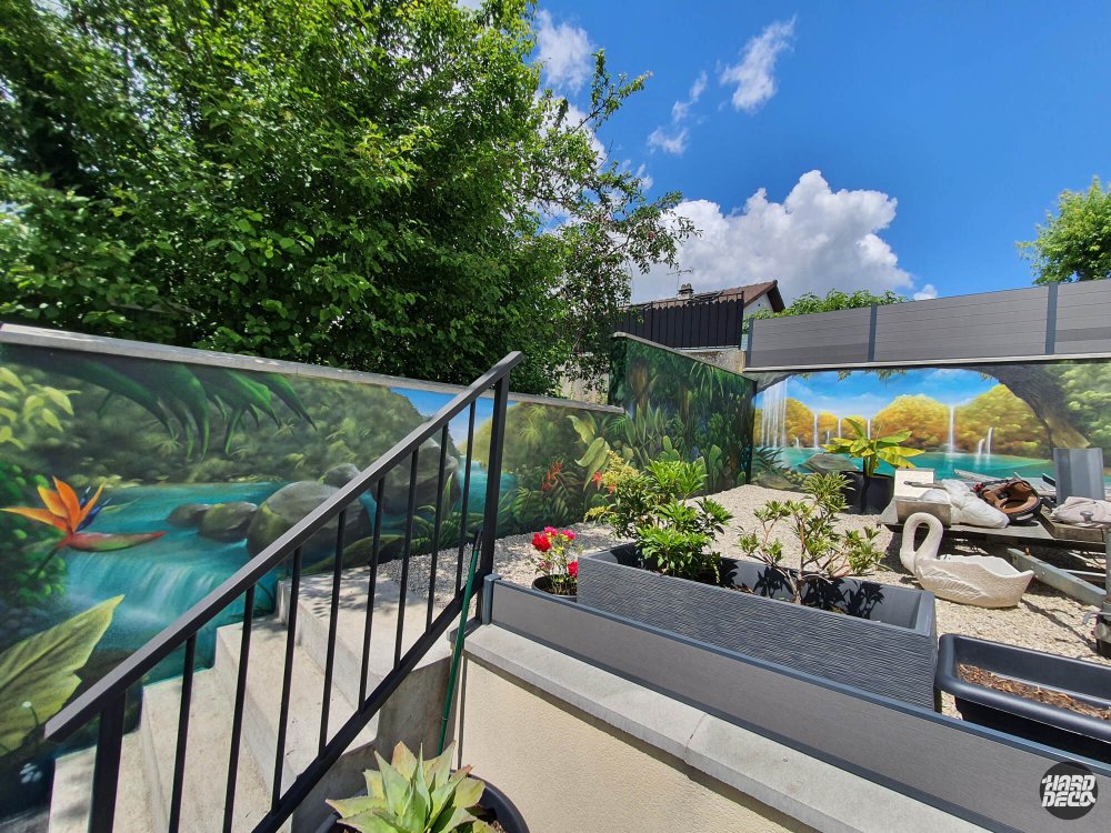 Fresque jungle tropicale sur deux pans de clôture en angle, vue depuis le jardin avec escalier et plantations