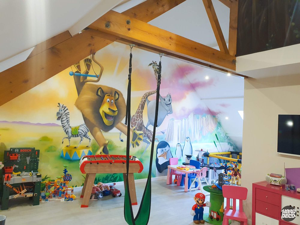 Vue d'ensemble salle de jeux avec fresque Madagascar sous charpente bois, lion et girafe peints à l'aérosol
