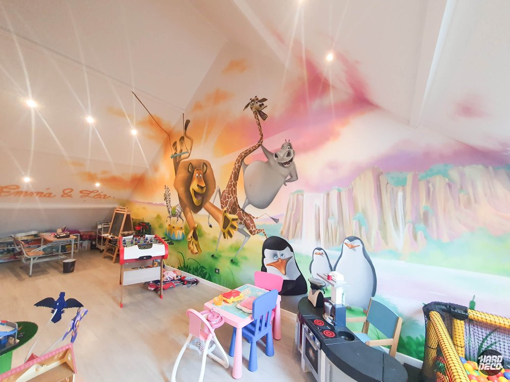 Fresque Madagascar salle de jeux sous combles : lion, girafe, pingouins et prénoms calligraphiés sur grand mur