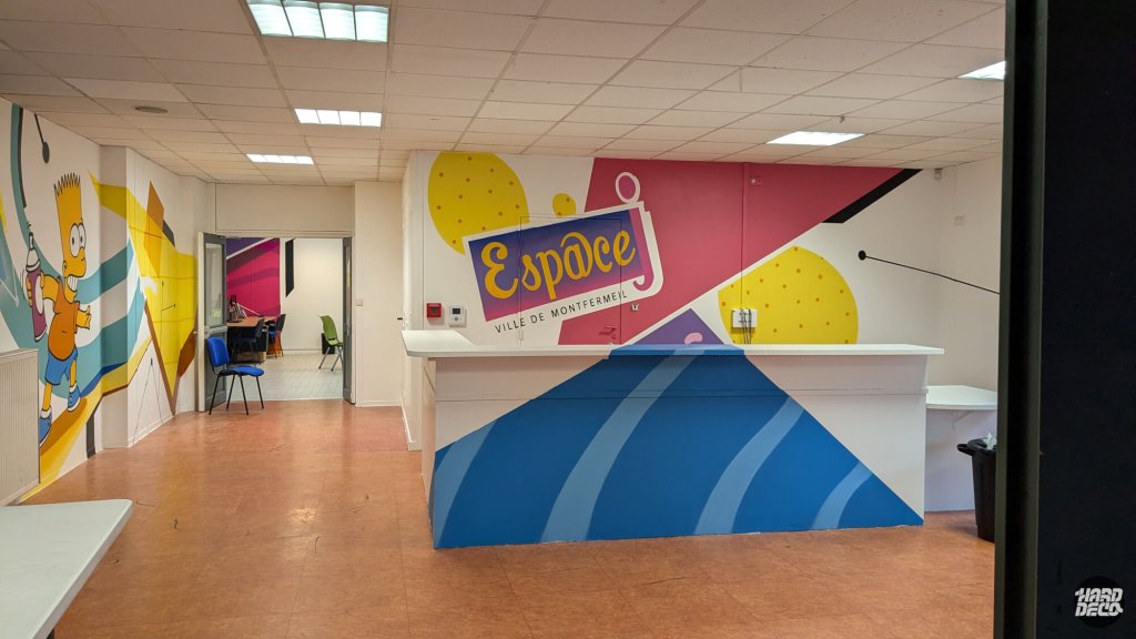 Logo Espace Ville de Montfermeil peint sur mur framboise et jaune, comptoir accueil bleu, décor mural collectivité