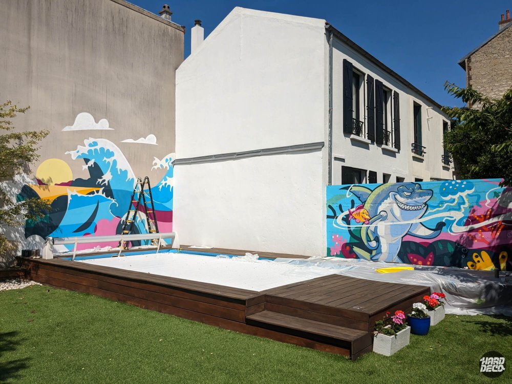 Jardin avec pelouse, piscine sur terrasse bois et deux fresques murales océan visibles sur les murs