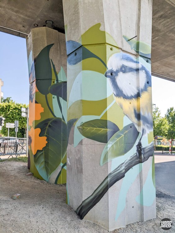Mésange bleue perchée sur branche, feuilles graphiques vert et doré sur pilier béton du RER A Torcy