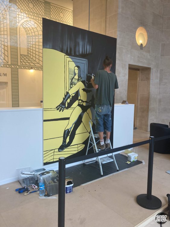 Artiste Samy au pinceau sur la toile Batman et Joconde en aplats jaune et noir, hall du Louvre