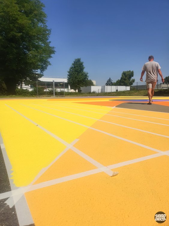 Phase de réalisation au rouleau sur terrain extérieur, aplat jaune vif et orange, artiste en cours de travail