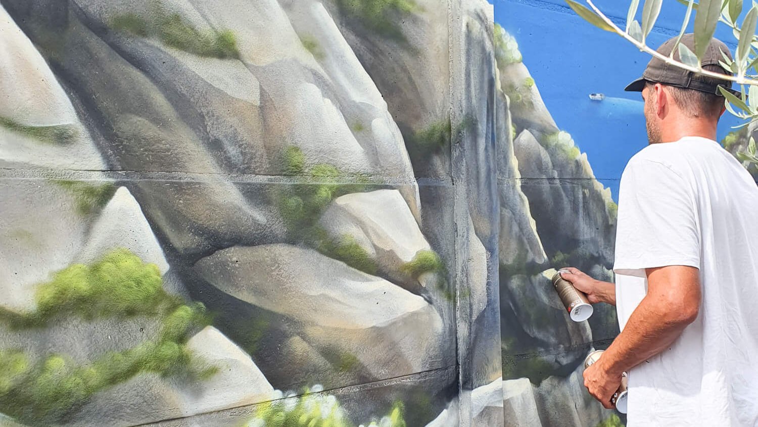 vue d'ensemble d'une fresque murale extérieure représentant un paysage avec arbres, rochers et ciel bleu, artiste en action