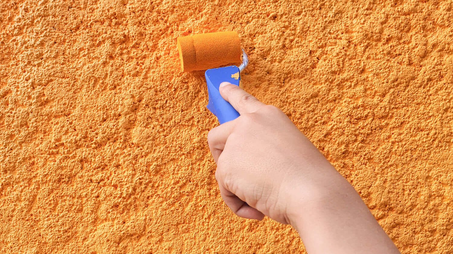 Main appliquant de la peinture orange avec un rouleau sur un mur à texture rugueuse